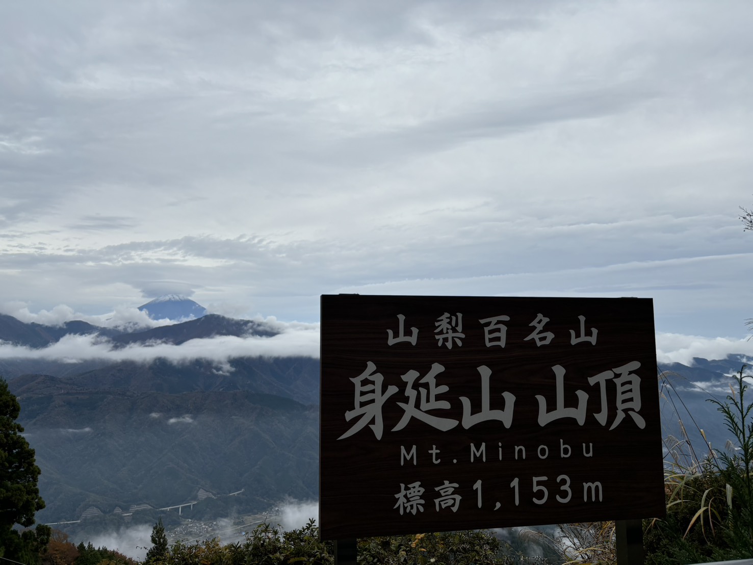 「山梨百名山 身延山山頂 標高1153m」の看板と、雲の上の富士山